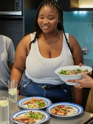 Três mulheres sorrindo preparam e servem pratos com salada e frango numa cozinha moderna, com copos e temperos na bancada.