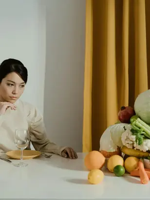 Mulher com paladar infantil ou seletividade alimentar sentada à mesa com expressão pensativa, cercada por pilha de vegetais coloridos.