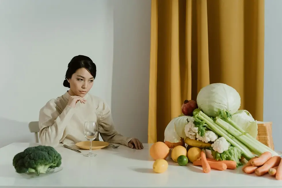 Mulher com paladar infantil ou seletividade alimentar sentada à mesa com expressão pensativa, cercada por pilha de vegetais coloridos.