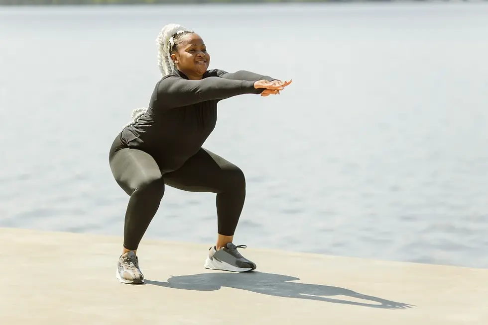 Mulher fazendo agachamento ao ar livre, com roupa de ginástica preta, perto de um lago. Dia ensolarado, expressão concentrada.