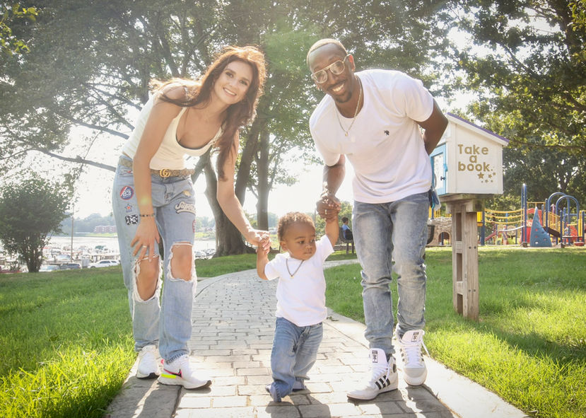 A smiling couple holding hands with their toddler son at a park on a sunny day, walking along a brick pathway.