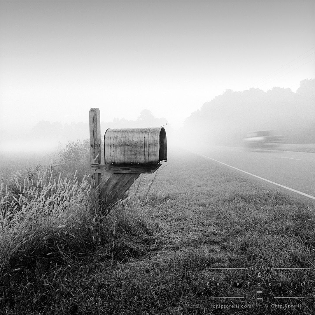 A rural mailbox at the side of a foggy road in perspective with a vehicle in motion speeding by in black and white.