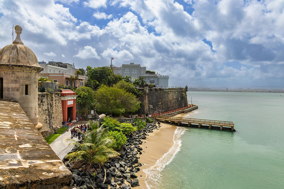 Historic fortress by the ocean, surrounded by lush greenery and rocky shore. Tourists walk near a red gate. Cloudy sky above.