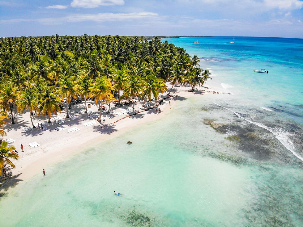Tropical beach with palm trees and lounge chairs. People walk and swim in clear turquoise water under a sunny sky. Boats visible offshore.