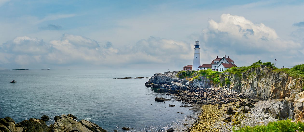 Lighthouse on rocky coastline with ocean view, green cliffs, and cloudy sky. White building with red roofs; serene and picturesque setting.