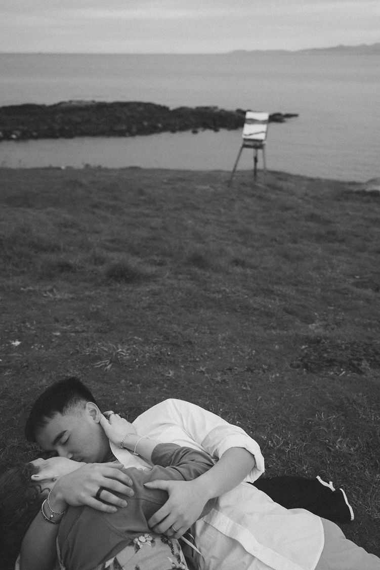 Couple kissing on a grassy cliff with an easel in the background in black and white