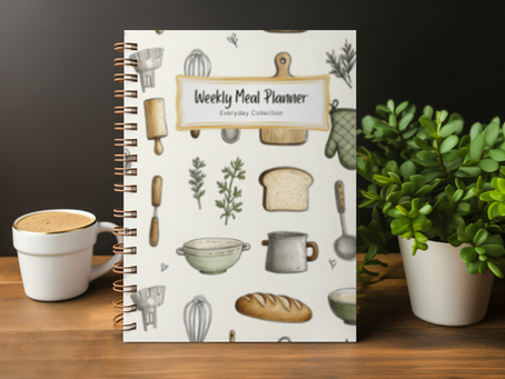 Spiral-bound weekly meal planner notebook on a wooden table with a coffee cup and plant, featuring a kitchen-themed cover design and grocery planning layout for weekly meal planning and organization.