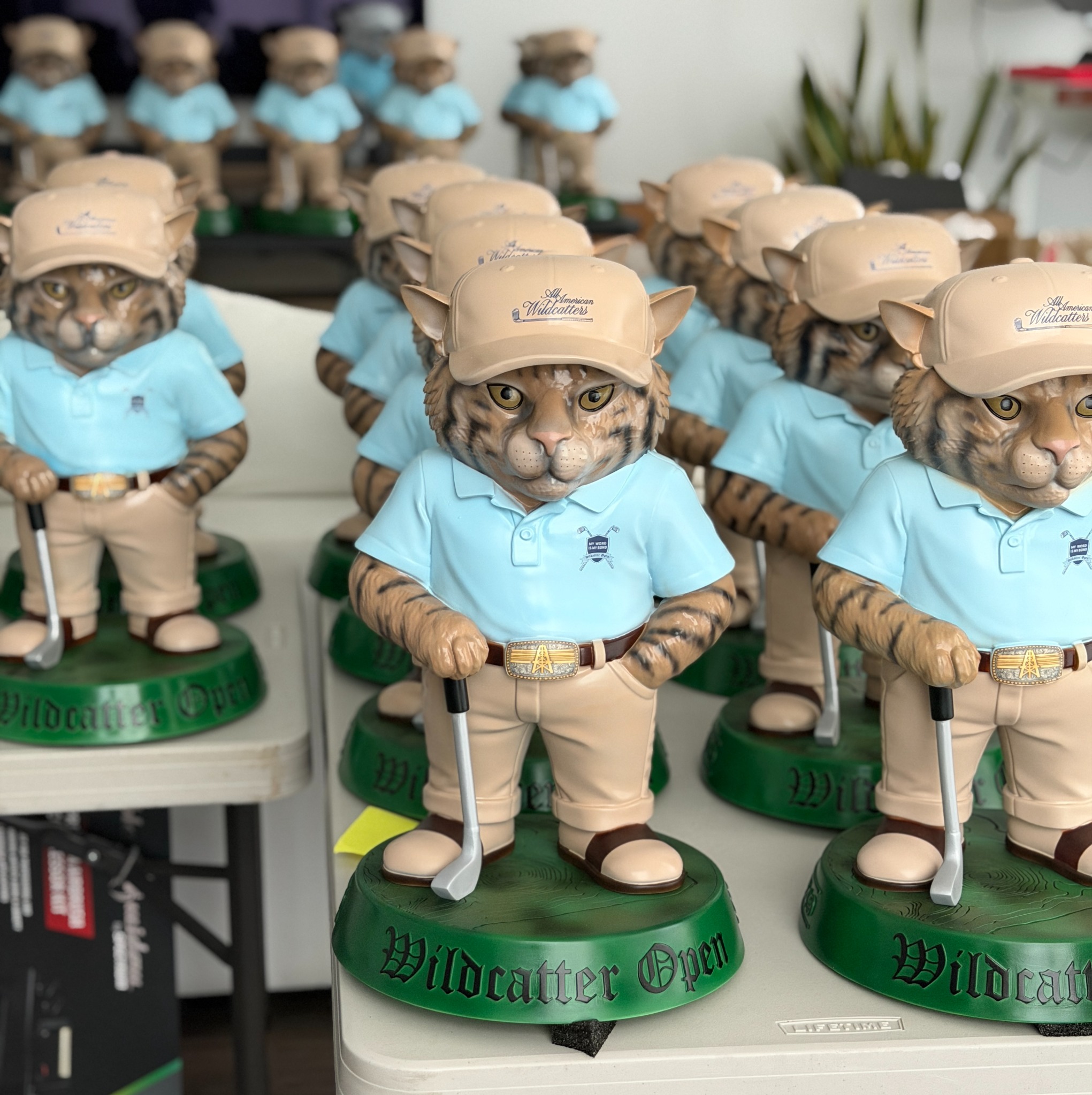 Rows of finished Wildcatter Open cat golfer figurines in blue shirts and tan caps on green bases