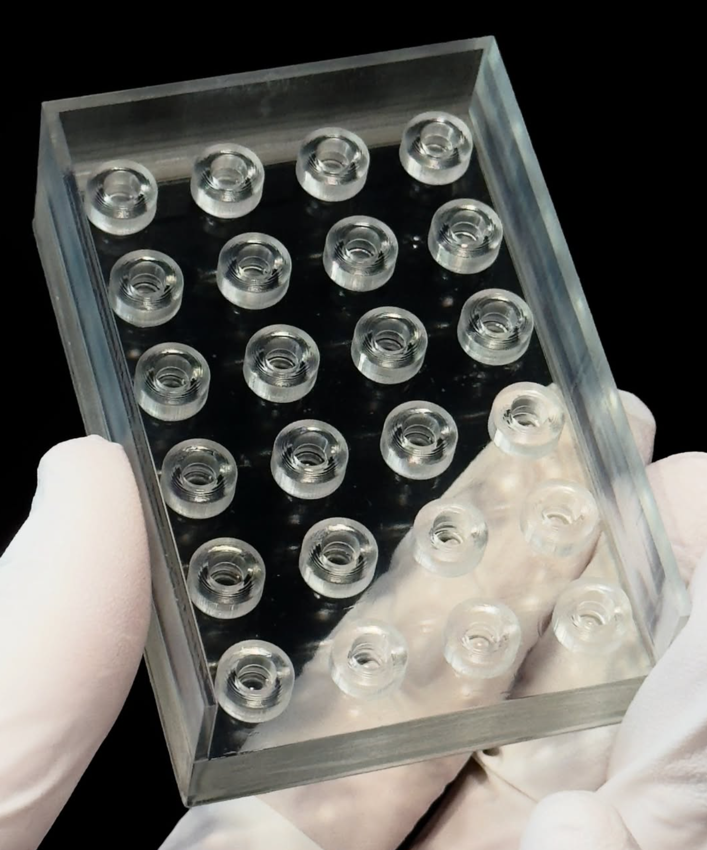 Gloved hands holding a clear resin rectangular component with an array of circular features
