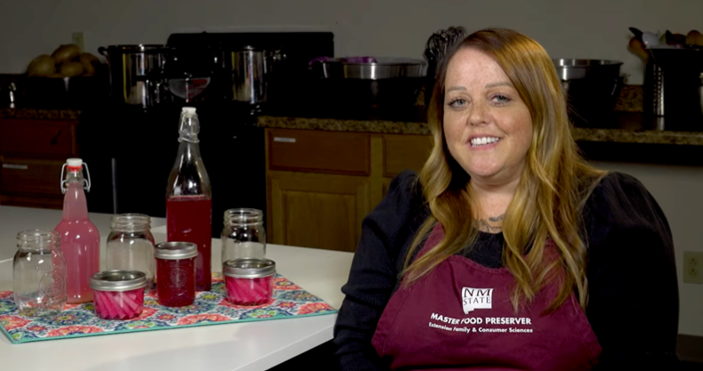 The image is a screenshot of the video discussed in the blog, portraying Amber, a master food preserver, next to a table with glasses and jars on top.