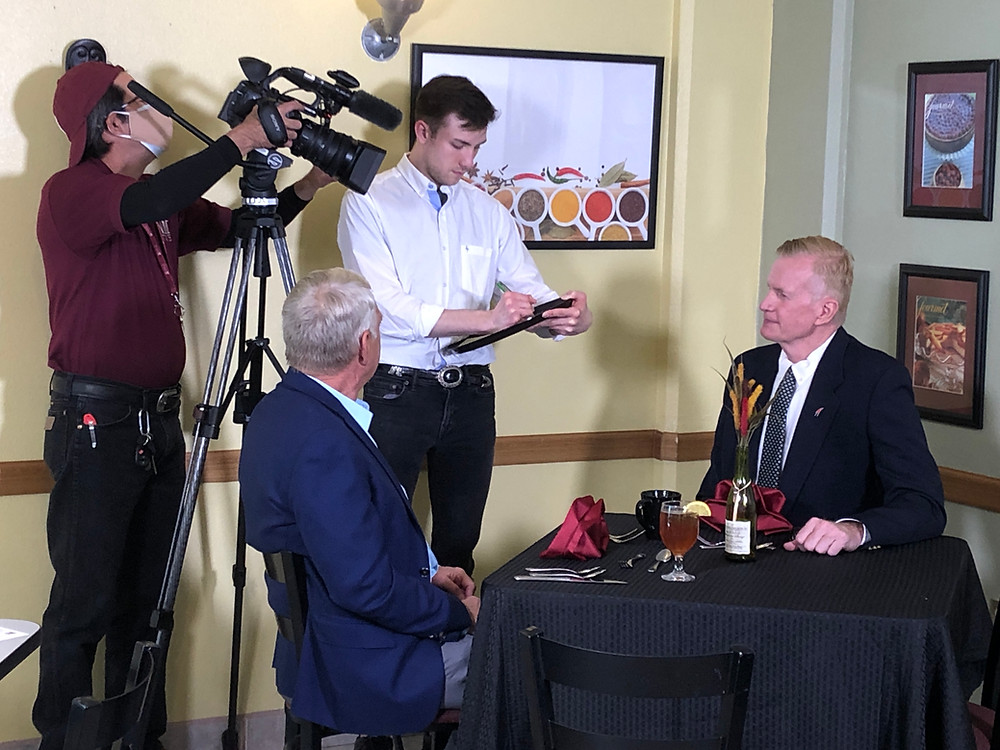 Two gentlemen sit at a table while a third person takes their order. This interaction is being filmed.