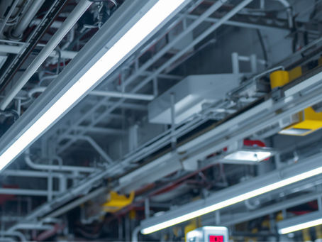 Teto industrial com tubos e luminárias de LED brancos. Estrutura metálica exposta com detalhes amarelos e fios aparentes. Ambiente moderno.