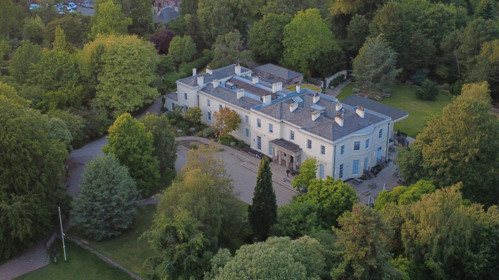 Aerial view of the Reader Manor House in Calderstones Park, Liverpool, showing a large white Georgian building surrounded by dense green trees, garden paths, and open lawns, captured during golden hour.