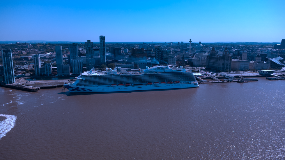 Aerial view of the Regal Princess cruise ship docked at Liverpool’s waterfront, with the city skyline and historic buildings in the background under a bright, clear blue sky.