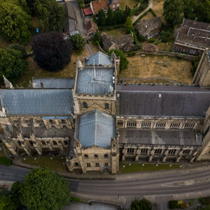 Aerial view of Ripon Cathedral showing its three square towers, intersecting roofs and surrounding trees beside a curving road.