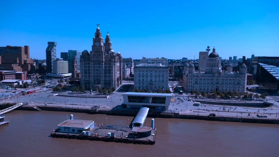 Aerial view of Liverpool’s Three Graces — Royal Liver Building, Cunard Building, and Port of Liverpool Building — along the waterfront with the Mersey Ferry terminal in the foreground and a clear blue sky above.