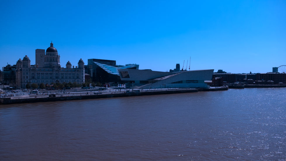 Aerial view of Liverpool’s waterfront showing the Port of Liverpool Building and the Museum of Liverpool beside the River Mersey on a clear sunny day, with people walking along the promenade.