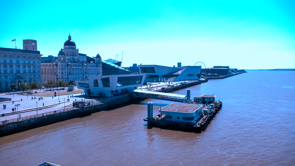 Aerial view of Liverpool’s Pier Head featuring the Port of Liverpool Building, Museum of Liverpool, Gerry Marsden Ferry Terminal, and River Mersey on a sunny day, with people walking along the waterfront promenade.