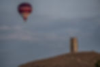 A photo of the red, black and white Haynes Motor Museum hot air balloon flying past Glastonbury Tor, showing St Michael's Tower and the top of the hill.