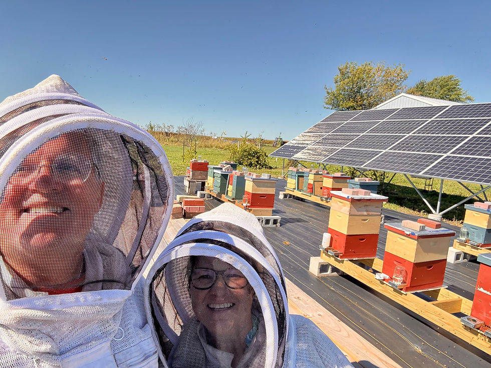 Two people in beekeeping suits smiling in front of bee hives and solar panels.