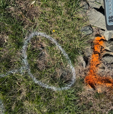 A white outlined circle and orange spray paint markings on grass, indicating underground utilities, with equipment visible in the background.