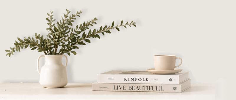 A plant in a vase, next to a cup of tea balanced on top of two luxury books.