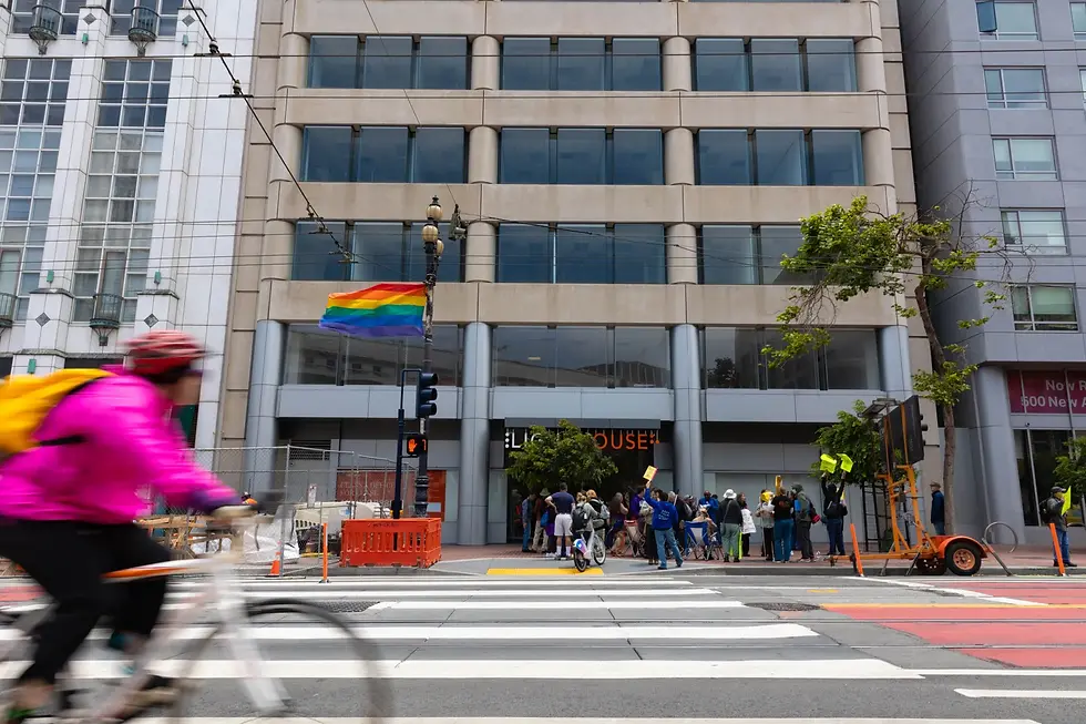 A cyclist in a bright pink jacket rides past a group of people holding signs outside the LightHouse for the Blind and Visually Impaired in San Francisco. A rainbow flag waves from a lamppost above the crosswalk. The building is gray with large windows and the word “LIGHTHOUSE” is partially visible above the entrance.