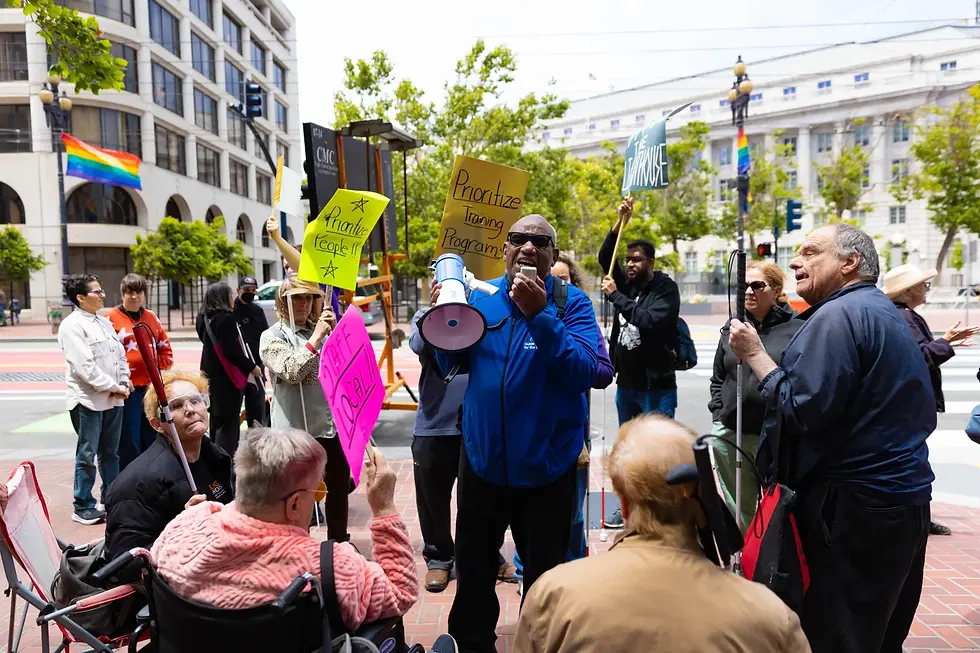 Anthony Lewis speaks into a megaphone during a June 12 protest in an urban setting. Protesters hold colorful signs, and a rainbow pride flag is visible in the background.