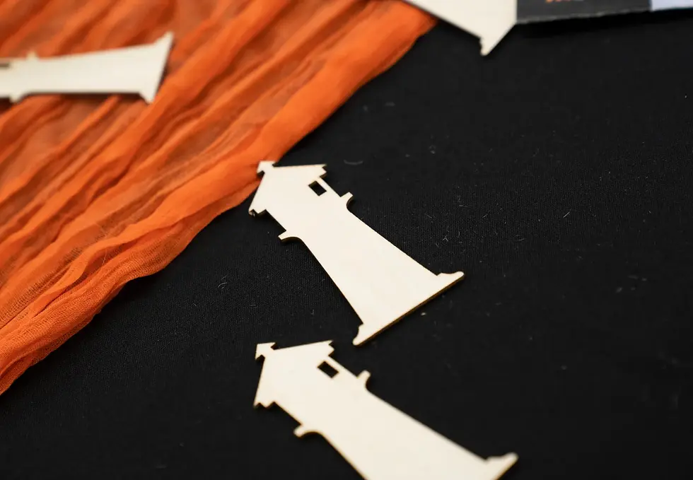 ID: Several thin, wooden cutouts in the shape of lighthouses placed on a black fabric surface. Two lighthouse shapes are clearly visible in the foreground, positioned diagonally, with simple details like the tower, roof, and small windows. An orange, wavy, sheer fabric runs along the top left portion of the image, adding a splash of color and texture. The overall aesthetic is tactile, crafty, and minimal, suggesting a handmade or decorative context.