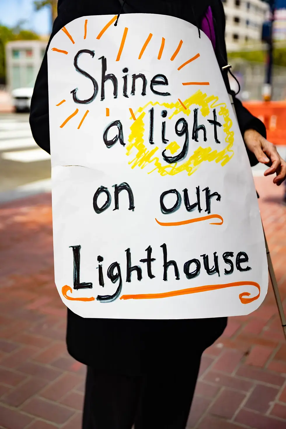 A hand-drawn sign reads “Shine a light on our Lighthouse,” decorated with a yellow sun and orange rays.