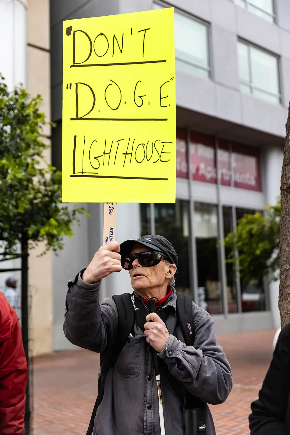 A man wearing dark sunglasses and a cap holds a bright yellow protest sign reading “Don't ‘D.O.G.E.’ Lighthouse.”