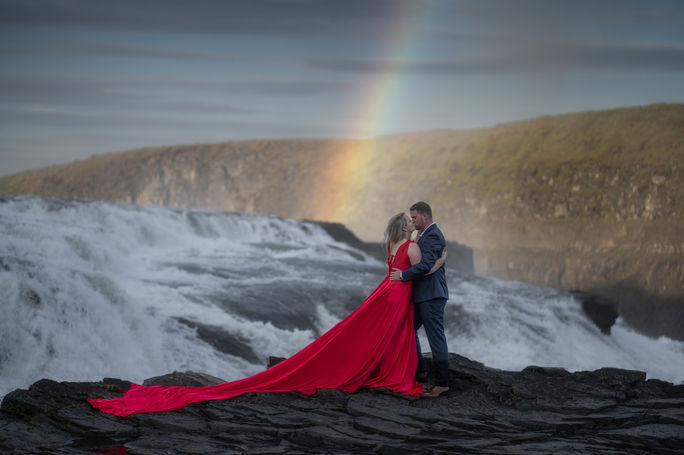 A bride-to-be in a red flowing dress poses with her partner at Gullfoss waterfall during their pre-wedding session. A beautiful rainbow arcs over them, enhancing the romantic and dramatic atmosphere of their shoot.