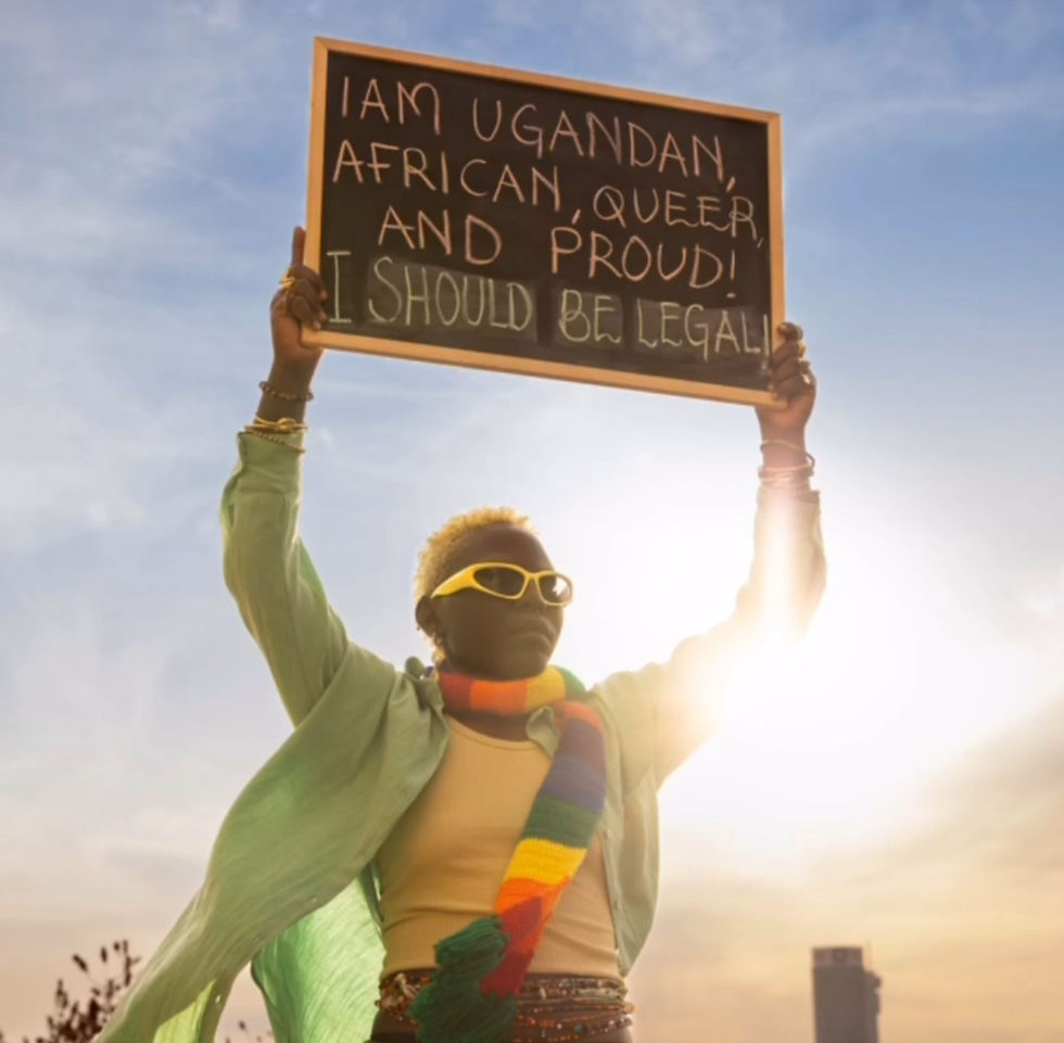 Papa De wearing green shades, a green t-shirt and and a rainbow-colored scart while holding a chalkboard which reads: "I am Ugandan, African, Queer and Proud! I should be legal."