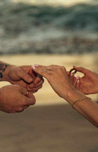 Detail shot of hands during the ring exchange, showing an engagement ring on the bride's hand and a wedding band being placed on a finger with the ocean blurred in the background.
