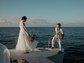 ride in a white tiered dress with a blue floral bouquet walking toward her groom on the deck of a private catamaran during an Oahu boat elopement