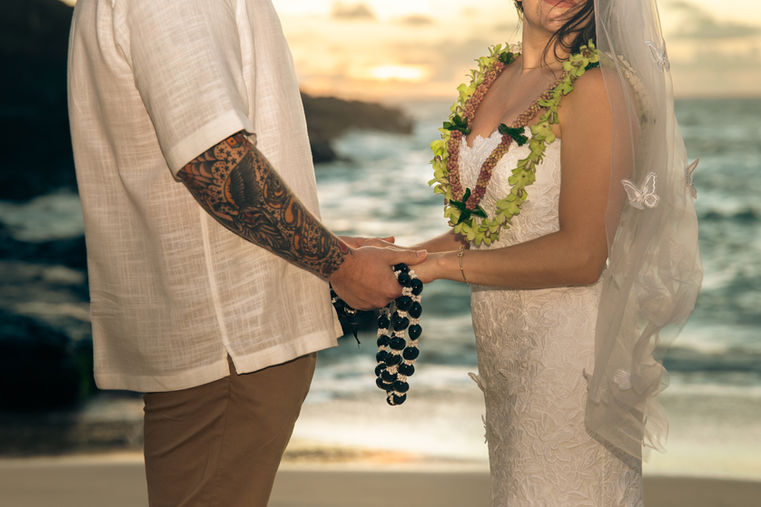 Close-up of a bride and groom holding hands and a dark beaded lei during their ceremony, highlighting the groom’s arm tattoo and the bride’s lace dress, green floral lei, and butterfly-embroidered veil.