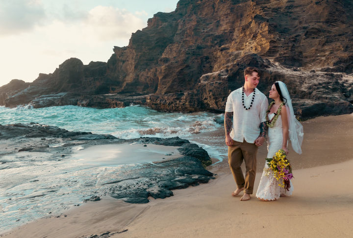 Bride and groom walking barefoot hand-in-hand along the sandy shore, flanked by crashing turquoise waves and steep cliff formations.