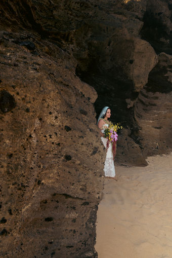 Bride framed by a massive textured rock face, standing in the sand holding her colorful tropical bouquet.