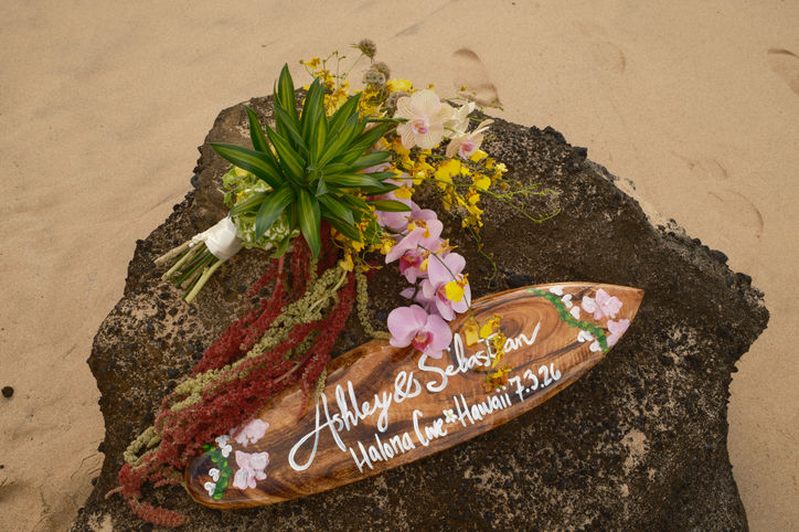 Top-down view resting on a dark rock, featuring a colorful tropical bouquet of pink orchids and yellow flowers alongside a personalized wooden surfboard keepsake sign reading "Ashley & Sebastian Halona Cove Hawaii 7.3.26".