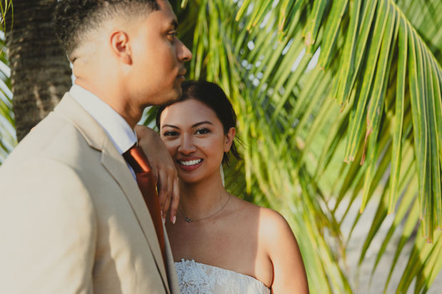 Close-up photo of Abriel smiling at the camera while leaning her arm affectionately on Darius’s shoulder outdoors under palm trees.