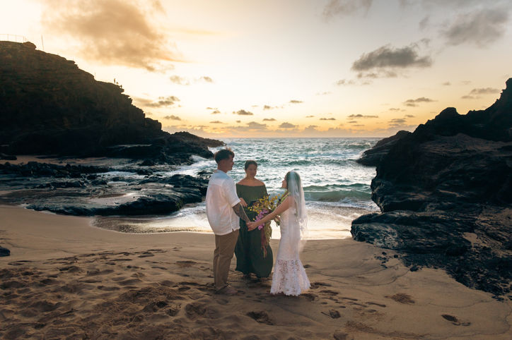 Wide view of a couple holding hands during an outdoor beach wedding ceremony at golden hour, with an officiant standing between them against a backdrop of ocean waves and rocky cliffs.