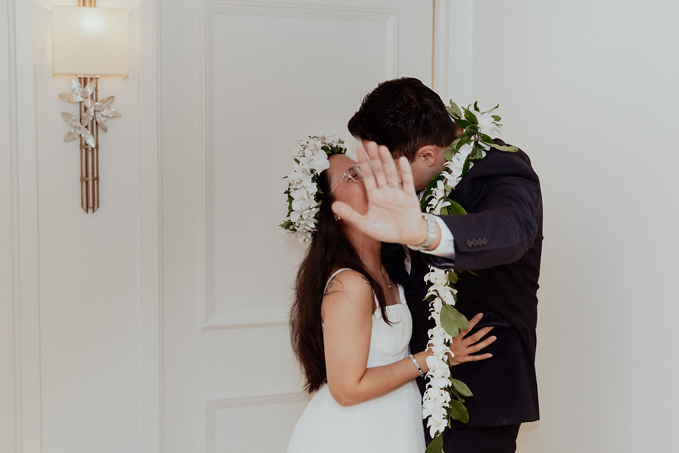 Playful bride wearing a flower crown and groom wearing a lei kissing while the groom holds up a hand to playfully block the camera lens.