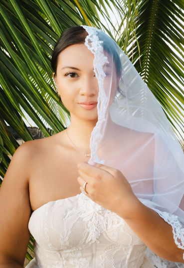 Portrait of Abriel holding up her lace bridal veil to partially cover her face with vibrant green palm leaves in the background.