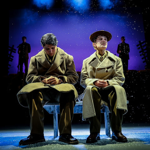 Two student actors in coats sit on a bench, looking thoughtfully on stage