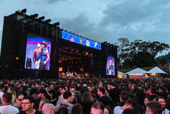 Visão ampla da multidão aproveitando o show em frente ao grande palco principal iluminado durante cobertura de festival de música ao ar livre.