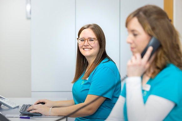 Szene an der Anmeldung: Im Fokus steht die Medizinische Fachangestellte Natalie Maurer, im vordergrund ist die Medizinische Fachangestellte Christina Schreiber verschwommen zu erkennen, wie sie gerade Telefoniert.
