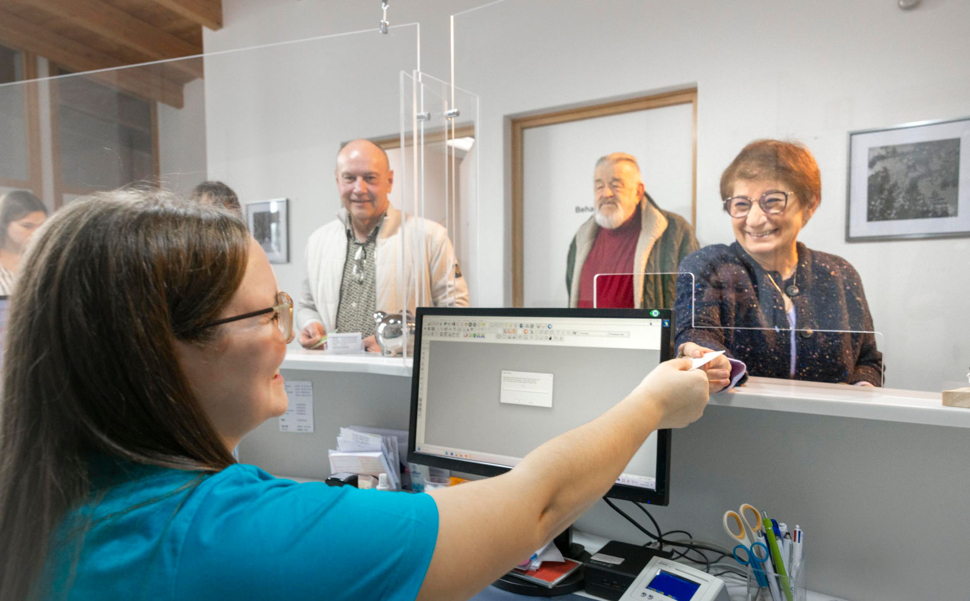 Eine Medizinische Fachangestellte nimmt an der Anmeldung die Versichertenkarte einer älteren Dame entgegen. Im Hintergrund warten noch zwei weitere Patienten. Alle Personen auf dem Foto lächeln