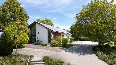 Außenansicht der Praxis. Das Haus ist umringt von vielen Planzen. Der Parkplatz liegt rechts vom Haus.
