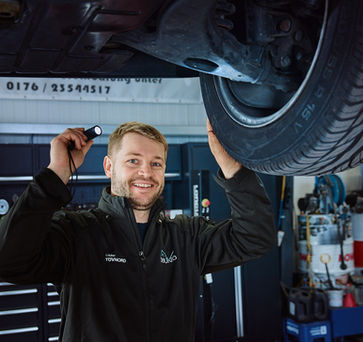 Thomas Huber inspiziert mit einer Taschenlampe den Boden eines Autos auf einer Hebebühne.