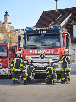 Vier Feuerwehrmänner stehen vor einem Feuerwehrauto und lächeln in die Kamera.